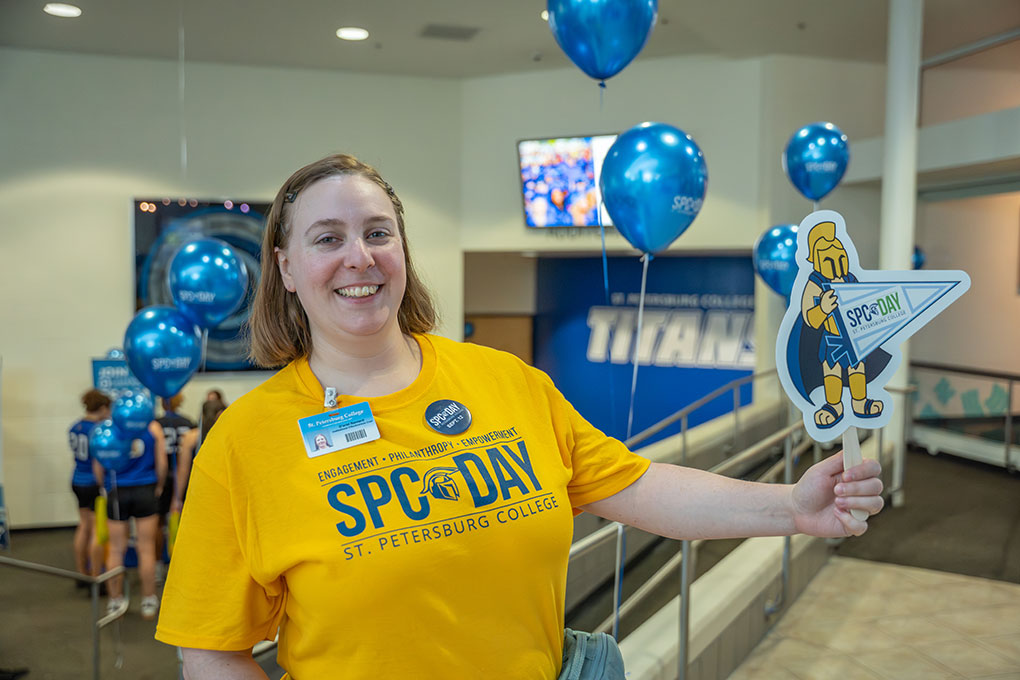 A woman at SPC Day 2025, smiling, wearing a yellow SPC Day shirt, and holding a hand fan picturing Titus the Titan waving an SPC Day pennant.