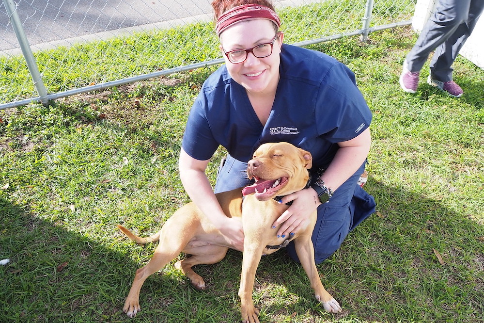 female student smiling holding onto a caramel colored dog with his tongue out