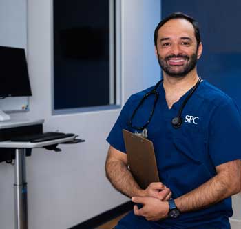 Uniformed nursing student in a medical room holding a clipboard smiling at the camera