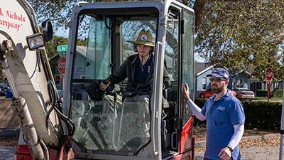 An SPC student wearing a straw operates an earth mover while being instructed by the owner wearing a blue cap