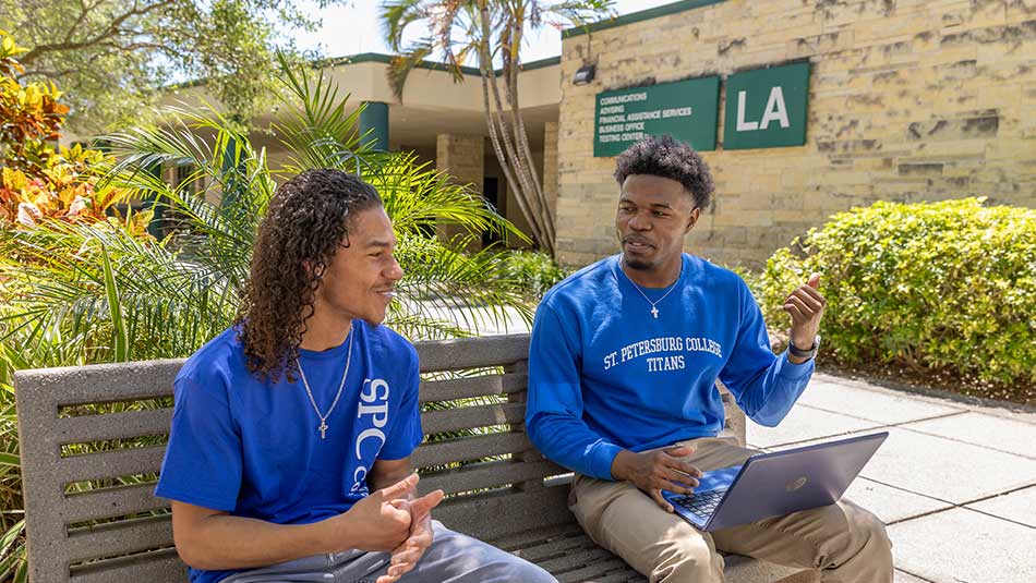 two male students in blue shirts talking while one holds a laptop a the clearwater campus
