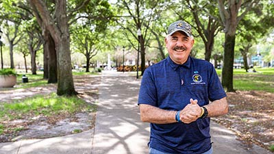 older male veteran wearing a camo baseball cap and a blue SPC polo shirt