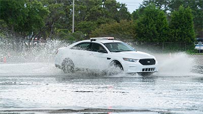 a white unmarked police training vehicle with red and white light bar on top driving through a gigantic puddle