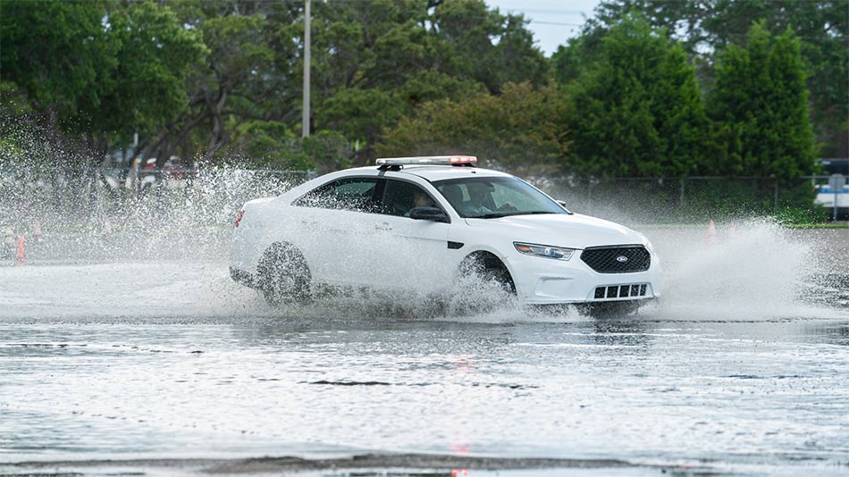a white unmarked police training vehicle with red and white light bar on top driving through a gigantic puddle