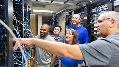 group of four students working in a network server room taking direction from their professor