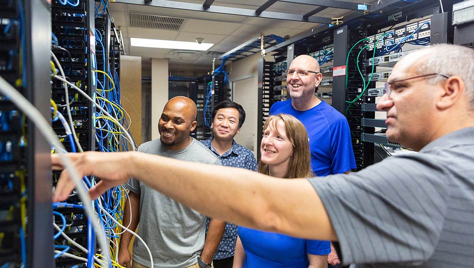 group of four students working in a network server room taking direction from their professor