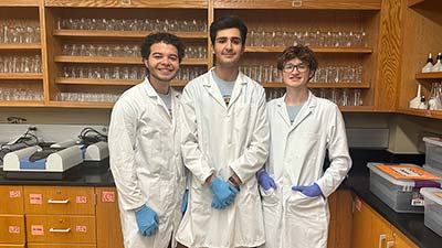 SPC students Vinicio Castillo, Eilya Yazdani and Nicholas Tsongranis wearing white lab coats smiling
