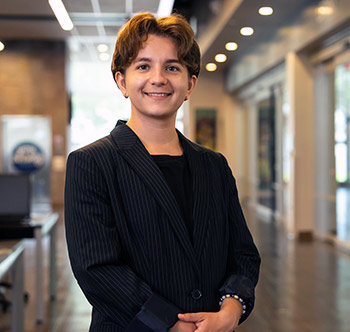 business student in a black suit with hands crossed standing in a hallway with windows