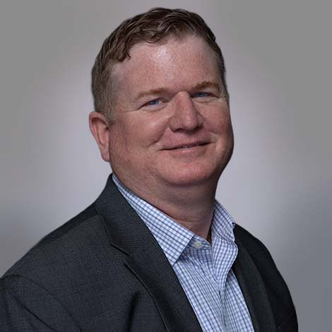 A portrait of Eric Abercrombie, wearing a gray suit coat over a white dress shirt with blue horizontal and vertical stripes.