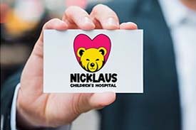 A hand displaying a card for the Nicklaus Children's Hospital with a golden bear face inside of a red heart shape.