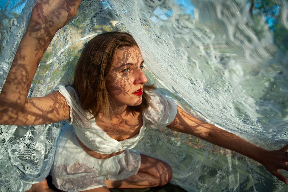 Student digital media project. Photograph of a woman crawling under a swath of white lace fabric. The lace patterning covers her in intricate shadows.