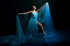 Student digital media project. Photograph of a dancer wearing a flowing blue dress and ballet pointe shoes. She stands in profile against a dramatic black background with her arms stretched outward and one leg extended with pointed toes. Gauzy blue material is draped around her shoulders and flows onto the ground like water.