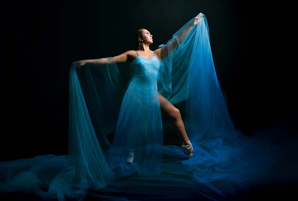 Student digital media project. Photograph of a dancer wearing a flowing blue dress and ballet pointe shoes. She stands in profile against a dramatic black background with her arms stretched outward and one leg extended with pointed toes. Gauzy blue material is draped around her shoulders and flows onto the ground like water.