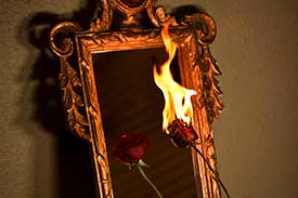 Student digital media project. Photograph of a hand holding a rose up to the reflective surface of an ornate mirror. In the reflection, the rose is untouched, while the one being held is on fire.