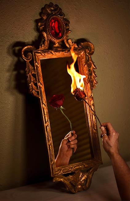 Student digital media project. Photograph of a hand holding a rose up to the reflective surface of an ornate mirror. In the reflection, the rose is untouched, while the one being held is on fire.