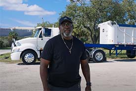 A truck driver, wearing a blue t-shirt and a baseball hat, standing in front of his white and blue dump truck. A truck driver, wearing a blue t-shirt and a baseball hat, standing in front of his white and blue dump truck.