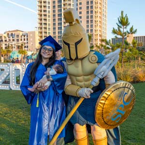 A new SPC graduate poses in her cap and gown, holding a baby, standing next to Titus the Titan