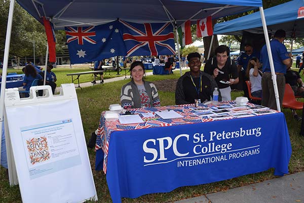 Three students working the SPC International Programs booth at an on-campus event, with Australian, British, and Canadian flags waving behind the tent canopy.