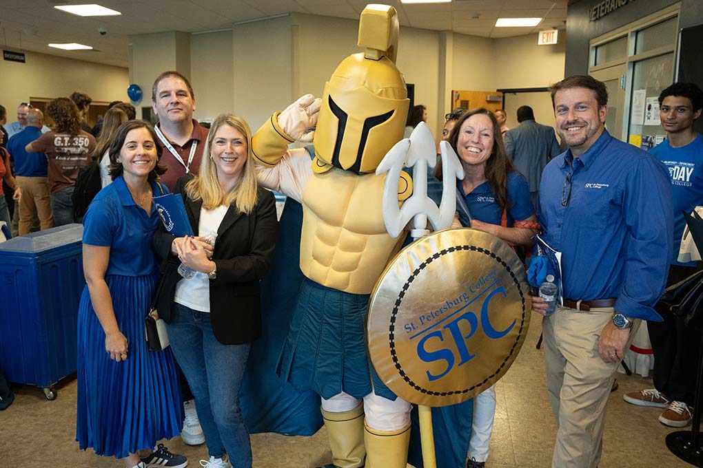 Five SPC staff posing with Titus the Titan at SPC Day 2024, with many people conversing in the background.