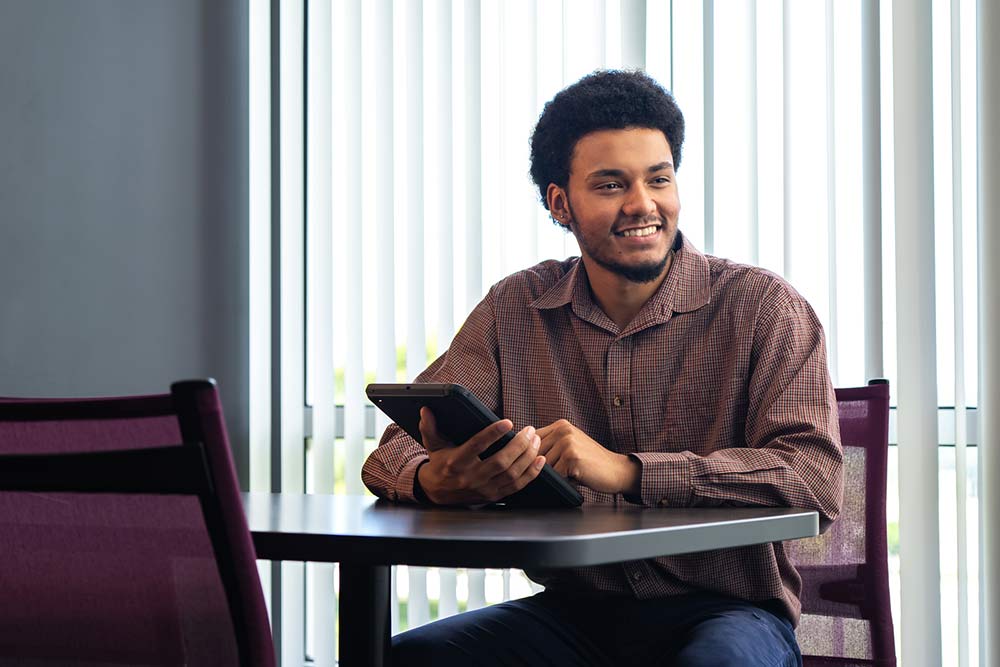 male business student wearing a brown shirt, smiling, with a tablet in his hand