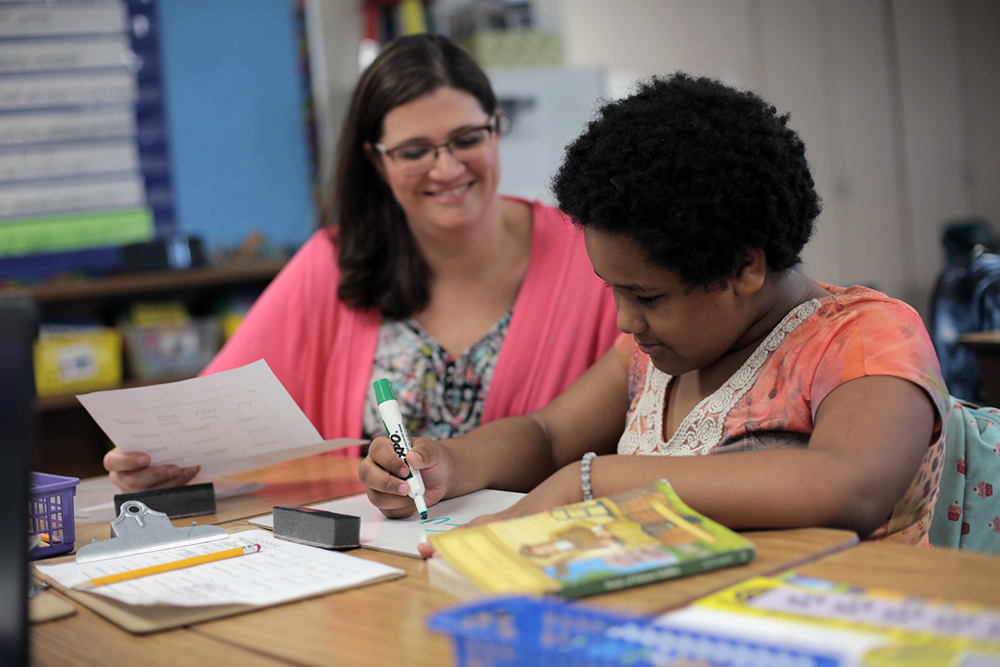 female teacher in a pink shirt sitting behind a desk next to a female student she is helping