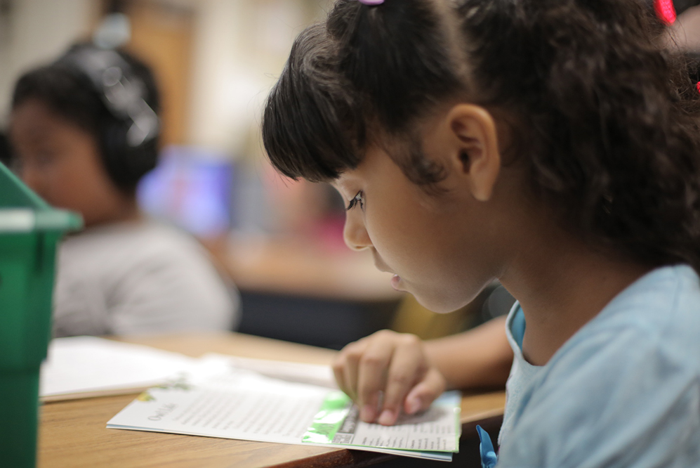 elementary aged girl with brown hair reading a book at a desk