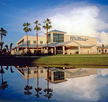 The SPC Seminole Community Library building behind 3 palm trees, under a blue sky and large, white cloud formation, and its mirror reflection in the water of the retention pond.