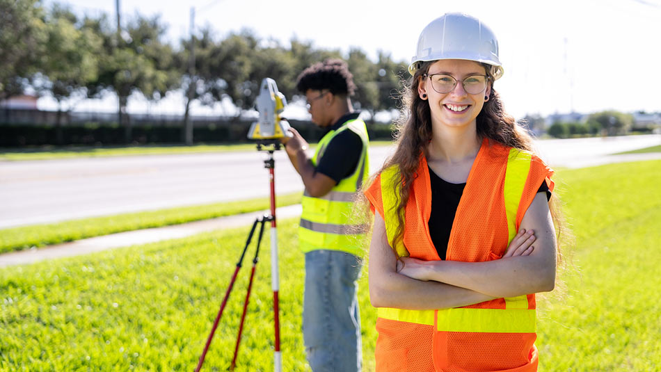 a male student using surveying equipment behind a female student wearing a hard hat and safety vest