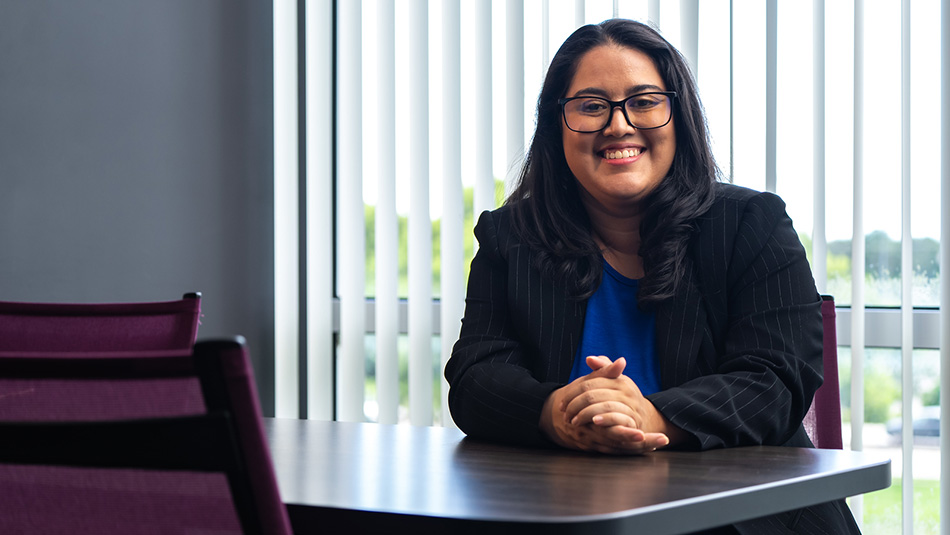 female student dressed in a blue shirt and black jacket sitting at a desk smiling