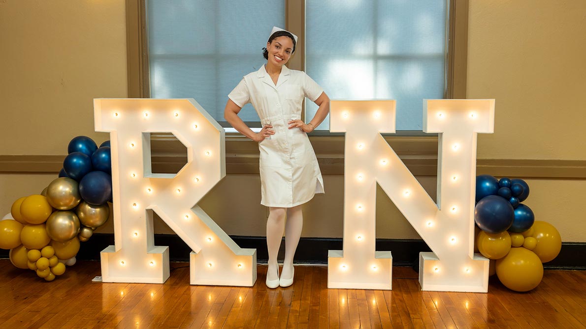 female nursing student in white nurse uniform standing in front of a light up sign that says RN