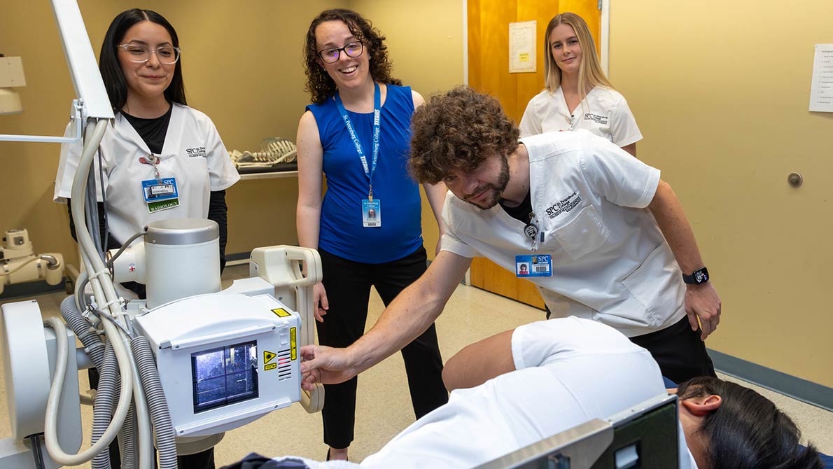 group of four radiography students in lab coats taking measurements of a fellow student on a table