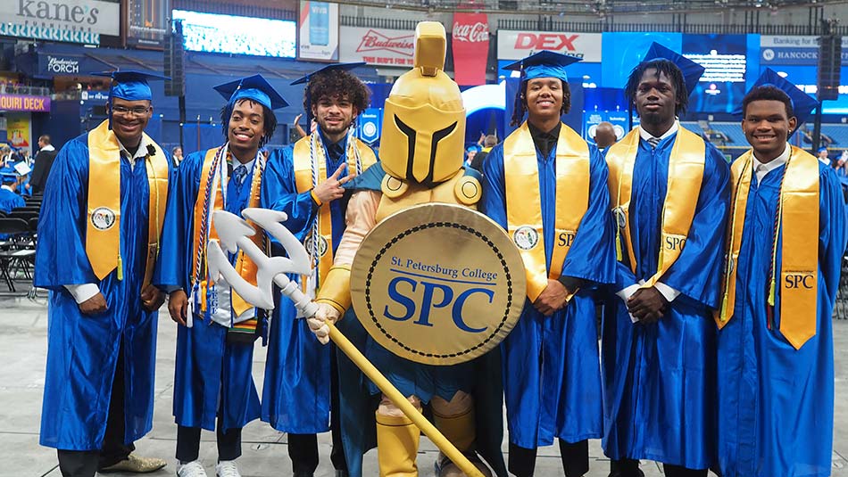 A group of 6 male graduates, wearing their caps and gowns, stands with Titus the Titan at the 2024 spring graduation ceremony at Tropicana Field.