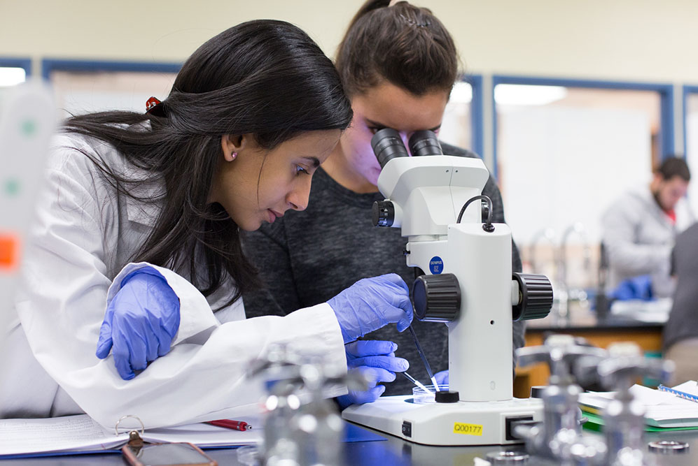two female SPC students in white lab coats peering through a microscope
